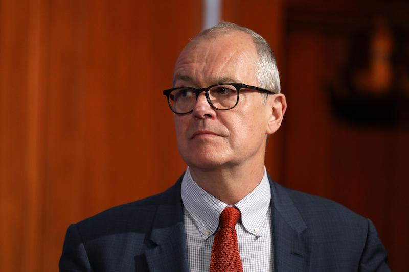 Sir Patrick Vallance in a grey suit and red tie, looking off to the left with a serious expression